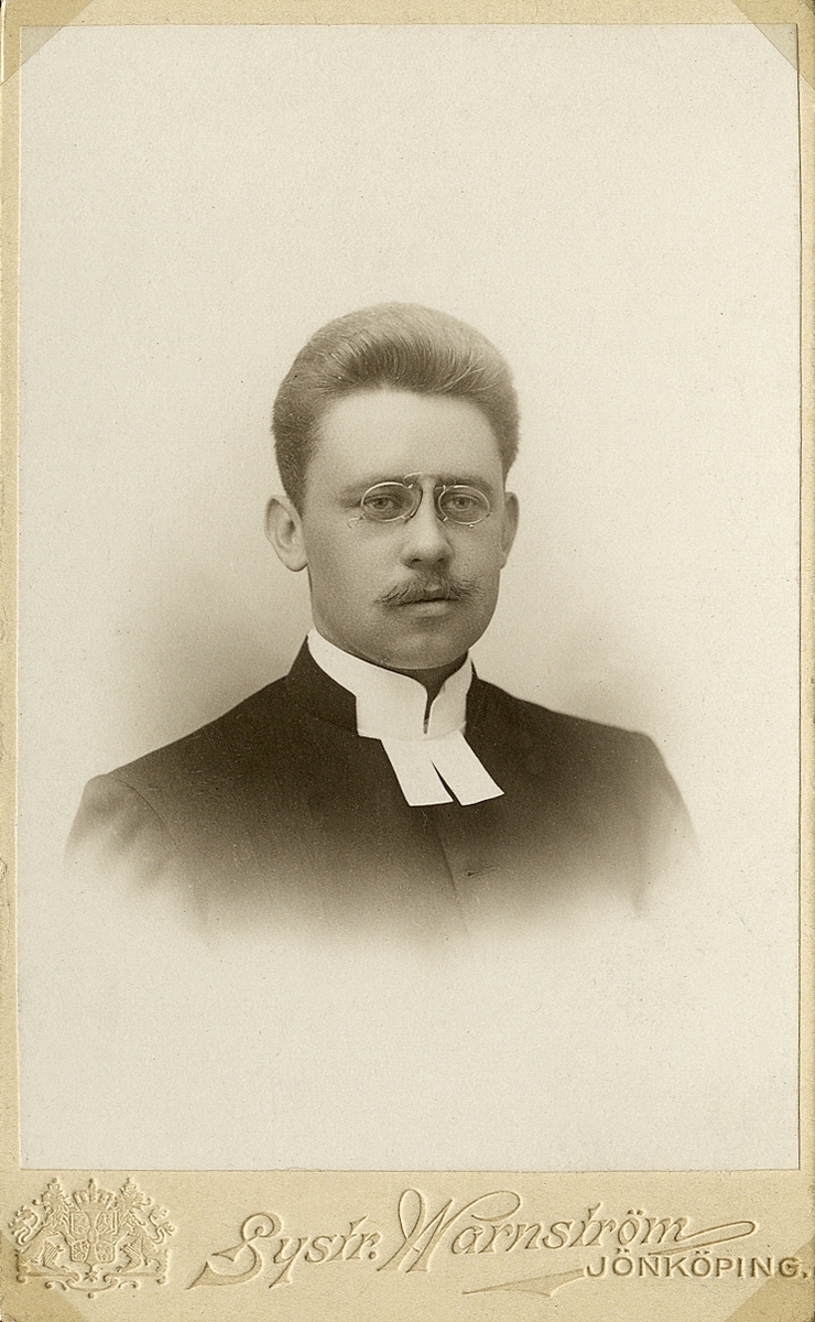 Foto av en man med mustascher och glasögon, klädd i prästrock och prästkrage.
Bröstbild, halvprofil. Ateljéfoto.