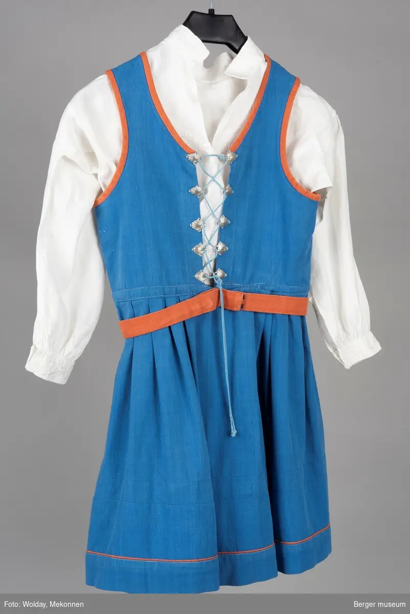 Barnebunaden/stakken er for jente i 10-12 års alderen. Den er blå med oransje detaljer, sølvhemper med skjellmønster, hvit bunadskjorte med hvite broderier. Hempene holder bryststykke sammen med en snor.