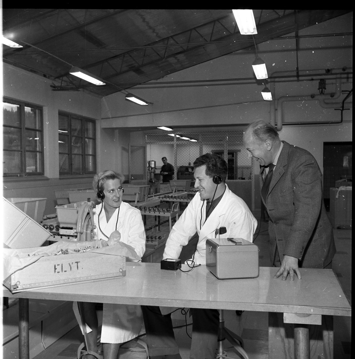 En kvinna och en man i vit arbetsrock sitter vid en arbetsstation på Rifa-fabriken i Gränna. Till höger står en man något lutad över dem. De skrattar.
De är från vänster Inga Johansson, Göran Andersson och G Eklund.