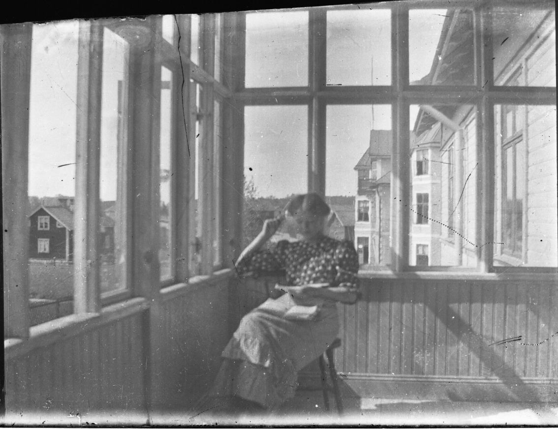 En okänd kvinna i prickig blus sitter på en solig inglasad veranda. Han har möjligen en bok i händerna och i knät.