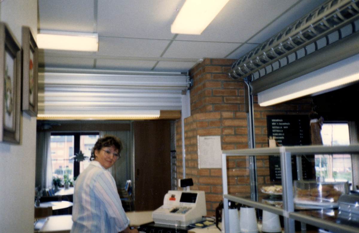 En kvinnlig personal står vid en kassaapparat i Brattåsgårdens matsal (på Streteredsvägen) cirka 1986 - 1990.