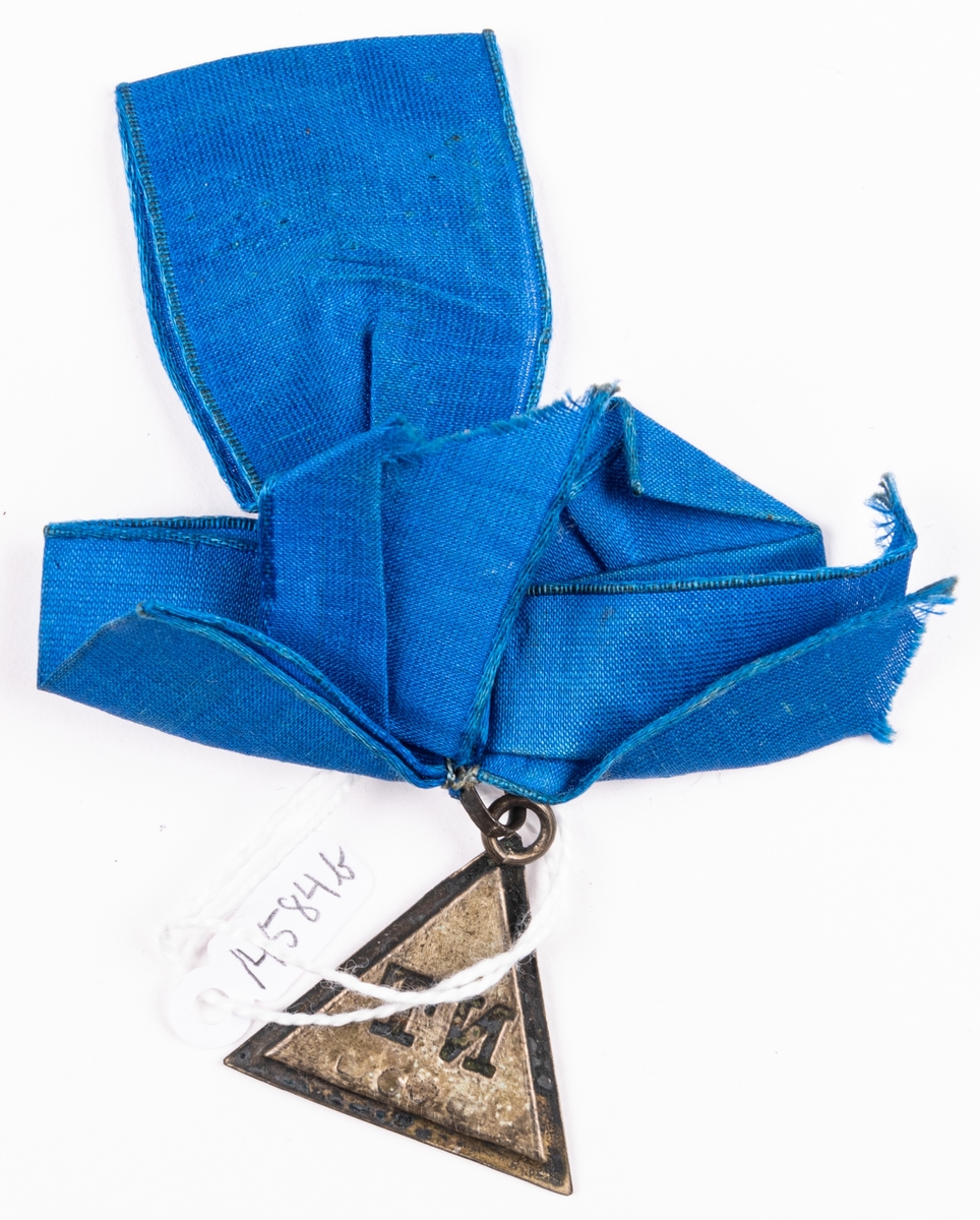 Triangelformat hänge i blå sidenrosett av siden.
Sällskapet NT. Stämplad.