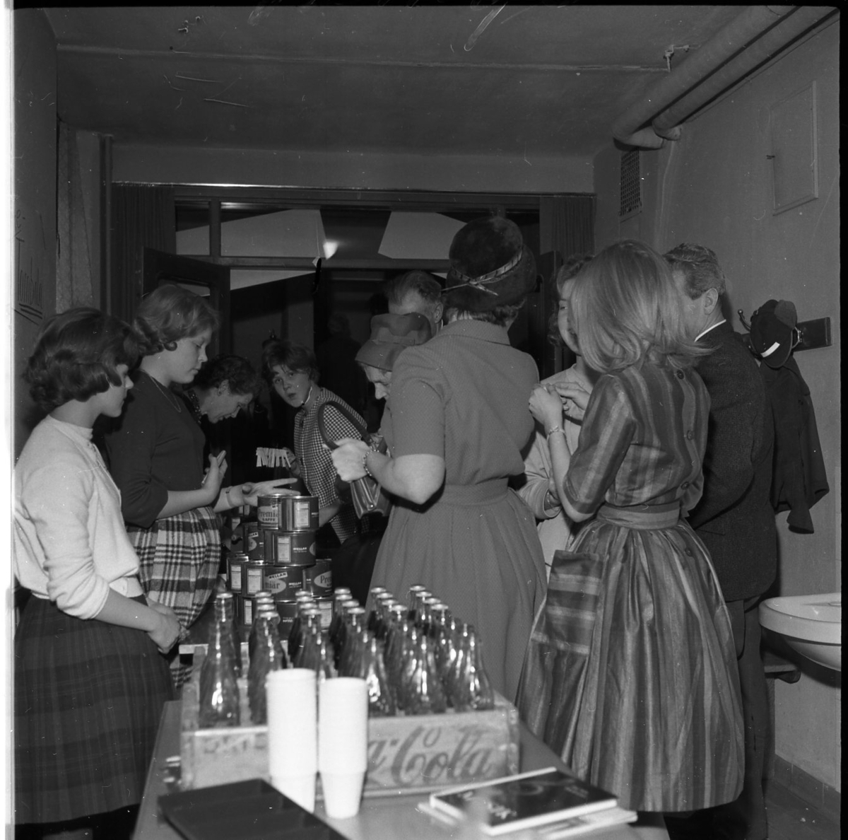 Unga kvinnor och enstaka män står i ett trång rum vid ett bord med läskedrycksflaskor (Coca-Cola) och konservburkar, det är lottvinster. Till vänster, bakom bordet, längst ned i bild står Birgit Heldebrandt. Till höger i randig klänning står Marita Wallmo.