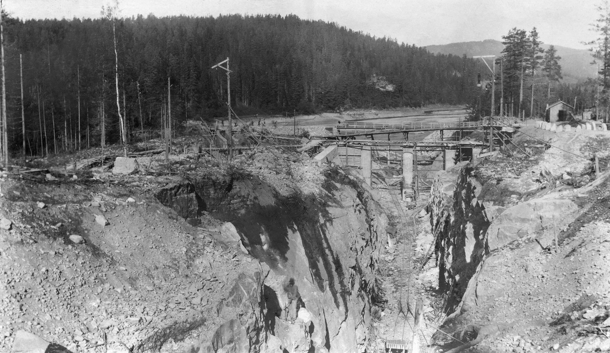 Anleggsarbeid i samband med utbygginga av Svelgfoss i Tinnåa, den østre delen av Skiensvassdraget, i 1906.  Fotografiet er tatt fra sør mot nord, med en utsprengt trasé med skinnegang for anleggsvogner i botnen.  Lengre framme ser vi at det er støpt kraftige betongsøyler for en damkonstruksjon på tvers av den utsprengte traseéen, og bak den igjen ser vi et par bru som forbinder østre og vestre del av anleggsområdet.  På vestsida skimter vi en del menn i arbeid på noe som synes å være en vegtrasé.  Her ser vi også at den øverste delen av tømmerrenna, som skulle ta fløtingsvirket effektivt forbi det kommende kraftverket, er ferdigstøpt.  I bakgrunnen ser vi Kloumannsjøen, som på dette tidspunktet var tappet svært lav, og den skogkledde Kvennåsen. 

For at tømmerkjøpernes og Skiensvassdragets Fellesfløtningsforenings interesser skulle holdes skadesløse i samband med denne utbygginga, ble det investert i ei 1 230 meter lang og en meter bred tømmerrenne, hvorav den øverste delen var støpt i betong.  Deretter fulgte en 450 meter lang tunnel sprengt i fjell med innlagt trerenne.  Ved utløpet av tunnelen var det et 30 meter langt skråplan, som førte vann og tømmer tilbake til elveleiet.  Anlegget hadde et fall på 1:100.  Poenget med disse konstruksjonene var å kunne føre tømmeret uskadd forbi kraftverket med minst mulig forbruk av vann.  I henhold til kontrakt med utbyggerne kunne Skiensvassdragets Fellesfløtningsforening disponere tre kubikkmeter vann per sekund i fløtingssesongen.  Med dette anlegget gikk fløtinga raskt og effektivt.  Nederst i Svelgfossrenna hadde tømmerstokkene en hastighet på 14 meter i sekundet. 