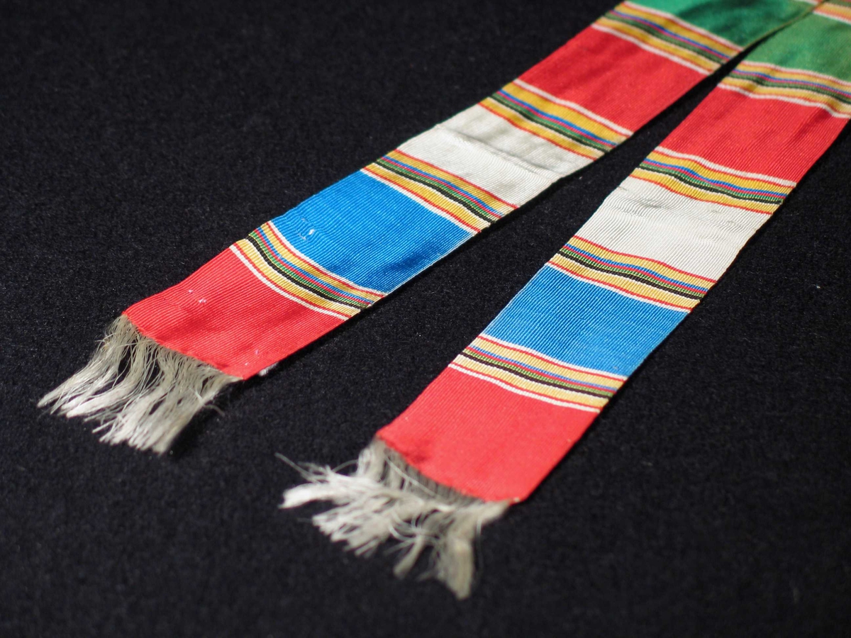 Tverrstripete silkebånd i fargene rødt, blått, hvitt og svart. Båndet har hvite frynser i begge ender som dannes av renningen.
