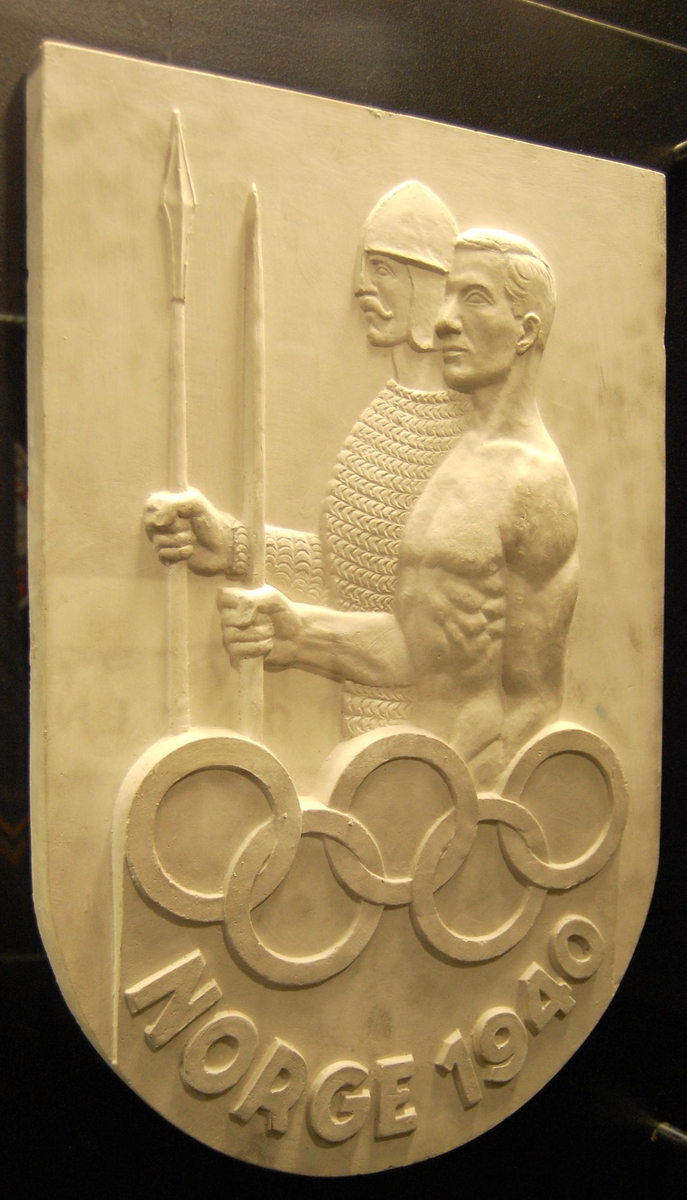 Skjoldformet plakett av gips med motiv av to menn, en i rustning og hjelm og en i bar overkropp. De olympiske ringene inngår i motivet.