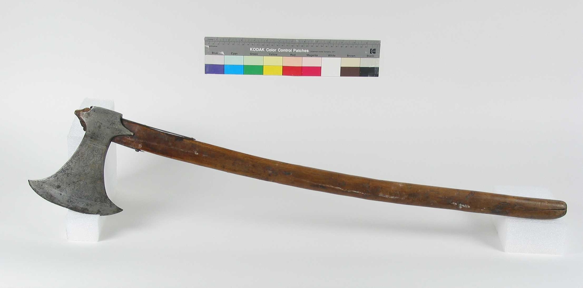 Symmetrisk blad. Bladet er tynt. 
Øsehammeren har skaftehull og fliker.
Etset dekor på begge sider. Stemplet TS på venstre side.
Gammelt treskaft med to orginale jernskinner med gravert dekor.
Jernskinnene er festet med nitter.