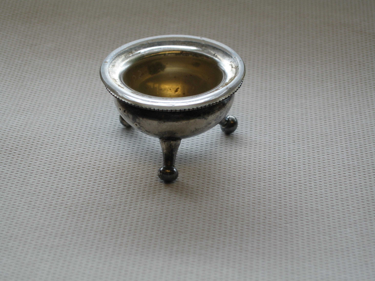 Saltkar av sølvplett og glass. Sølvfoten formet som en gryte med tre bein. Halvkuleformet glassinnlegg med utbrettet kant.