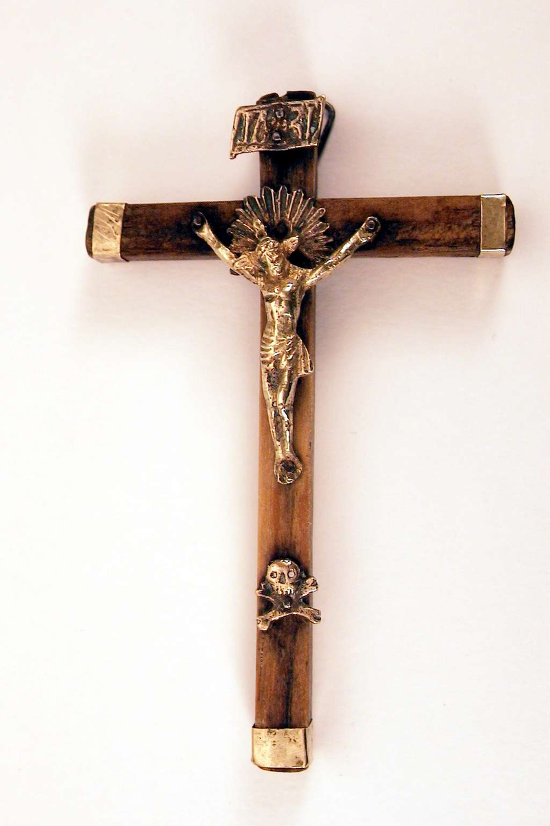 Kors av tre, med endebeslag i messing. Kristusfigur og dødningehode er i støpt messing. Over kristusfiguren en messingplate med bokstavene I N R I. Oppheng i metall på baksiden.

