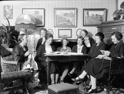 Interiør. Åtte kvinner rundt et bord. Malerier.