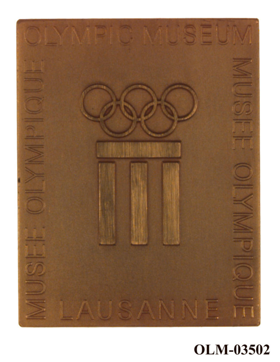 Gullfarget medalje med motiv av en gresk bygning med tre søyler.