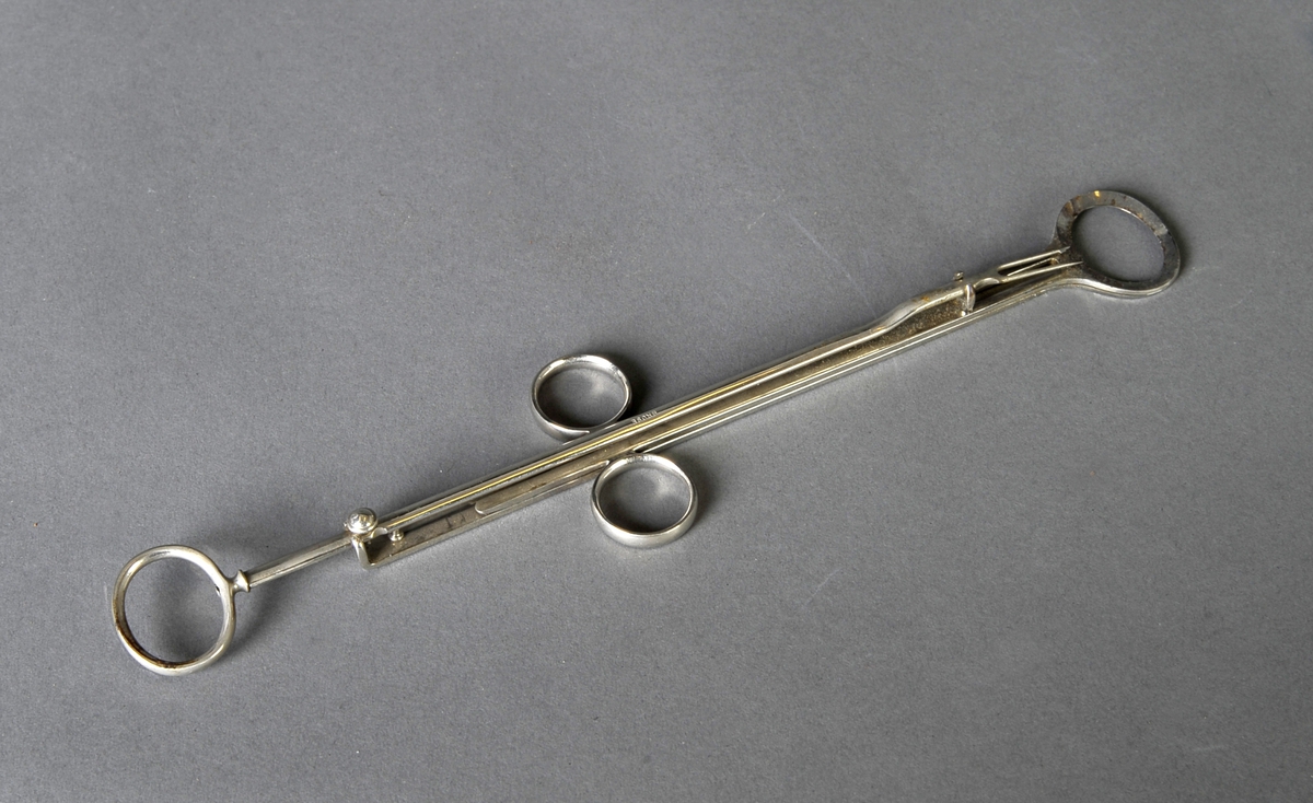 Metallstang som ender i 2 spisser med mothake, to flate metallstenger som ligger mot hverandre og ender i hver sin ovale kniv. 3 ringer til fingertak.
