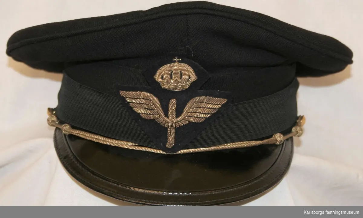 Storlek: 56
Skärmmössa m/1930 av mörkblått tyg mössband av svart räfflat silke. Svart lackerad skärm, försedd med hakrem av guldsnodd fäst vid två 14 mm uniformsknappar samt flygemblem.