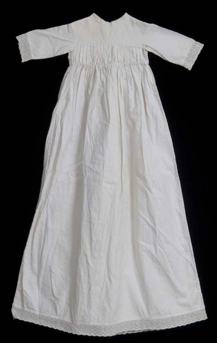 Hvit dåpskjole med flate folder (midtfold) i midja. Blonder på armlinningene og nederst på kjolen. Knyting bak. Hører sammen med lue (dåpslue)  NF.2007-0691A. 
