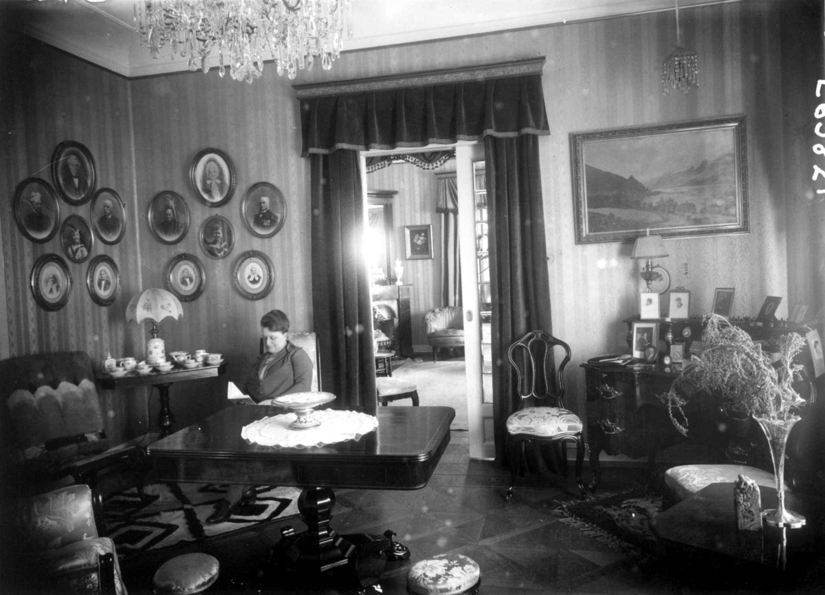 Trapp-Meyer. Stueinteriør 1925. Stoler og bord. Kvinne med bok. Portretter og maleri på veggene. Lysekrone. 