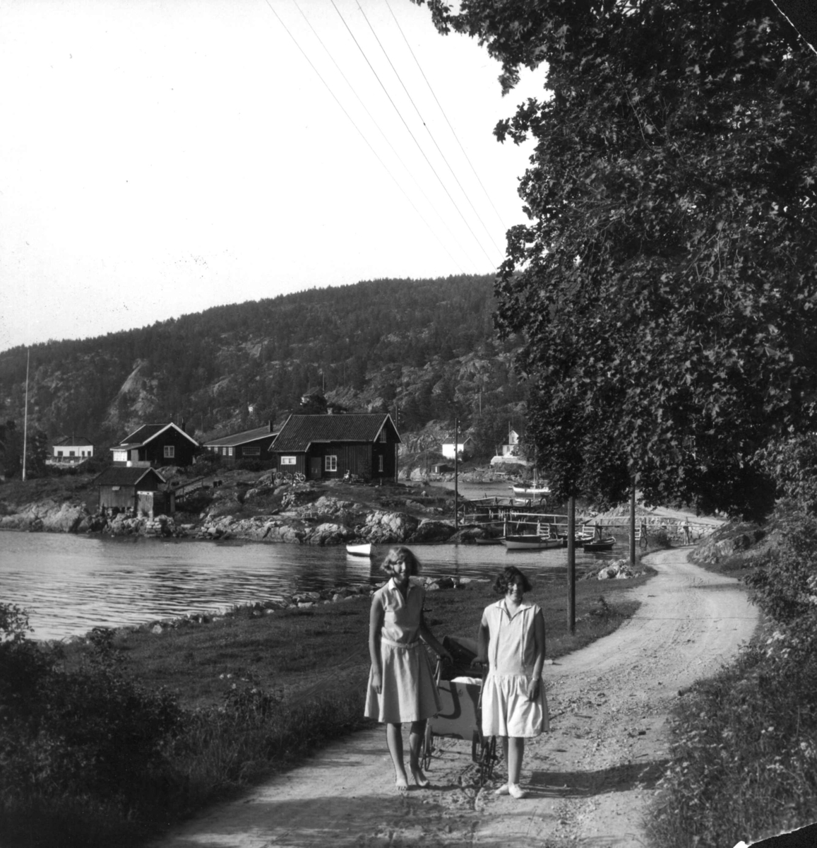 Drøbak, Akershus 1929. To unge kvinner i sommerkjoler med barnevogn står på en vei som går langs sjøen. Bebyggelse og båter ses i bakgrunnen.