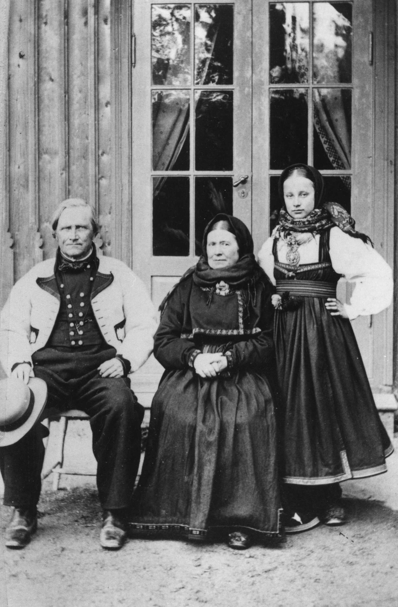 Portrett, gruppe i drakter fra Aust-Telemark, eldre mann og kvinne, jente. De poserer foran dør til bolig. Mannen holder en hatt i hånden.
Visittkortformat.