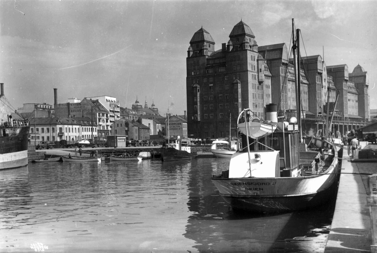 Havnelageret, Oslo 1937. Sett fra sjøen. Båter ved kai. Bebyggelse i bakgrunnen. Gate med biler og fotgjengere.