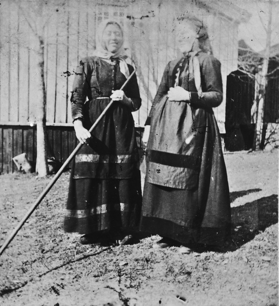 Kvinner på gårdstunet, Lunden gård, Hegglandsgrend, Fyresdal, Telemark, med stabburet i bakgrunnen. Kvinnen til høyre antas å være Kristi O. Haugen født 1850, fra nabogården nedre Haugen, et tun som i dag er slettet. Hun utvandret til USA i 1890 og kan ha blitt fotografert i forbindelse med avreisen. Kvinnen til venstre er antatt tjenestejenta Sigrid T. Toen (1876-1936) med skaut og rive.  Hun var en av flere barn av Torjus Auverson N. Toen (1850-1920), gift 1870 med Hege Halvorsdatter Geitestad (1850-1938) frå Rusto/Jordet i Valle. De var innerster og husfolk og bodde ulike steder omkring Lunden. Sigrid ble gift 1903 på Tveiten i Hauggrend.
Innsendt kommentar: "Ho med riva var tydeleg ei tenestegjente, ho hadde ikkje "tullplagg", men skaut. Ikkje knappar framme. Snipp på skautet fram som då vart litt meir til pynt. Ho måtte ha høg status på garden då ho vart fotografera i tunet."