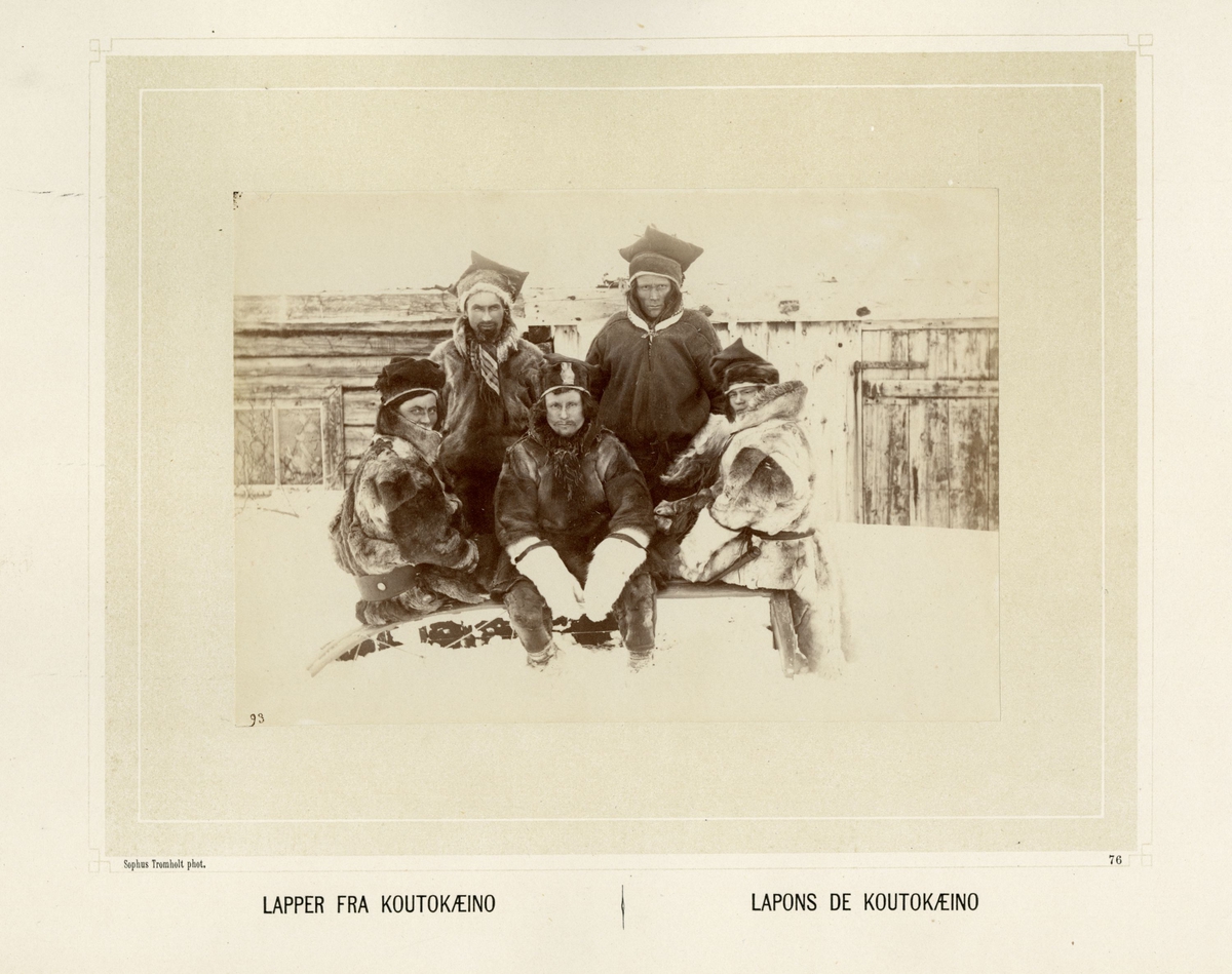 Gruppe med samer fra Kautokeino, sitter på en pulk i snøen kledd i pesk og skaller.