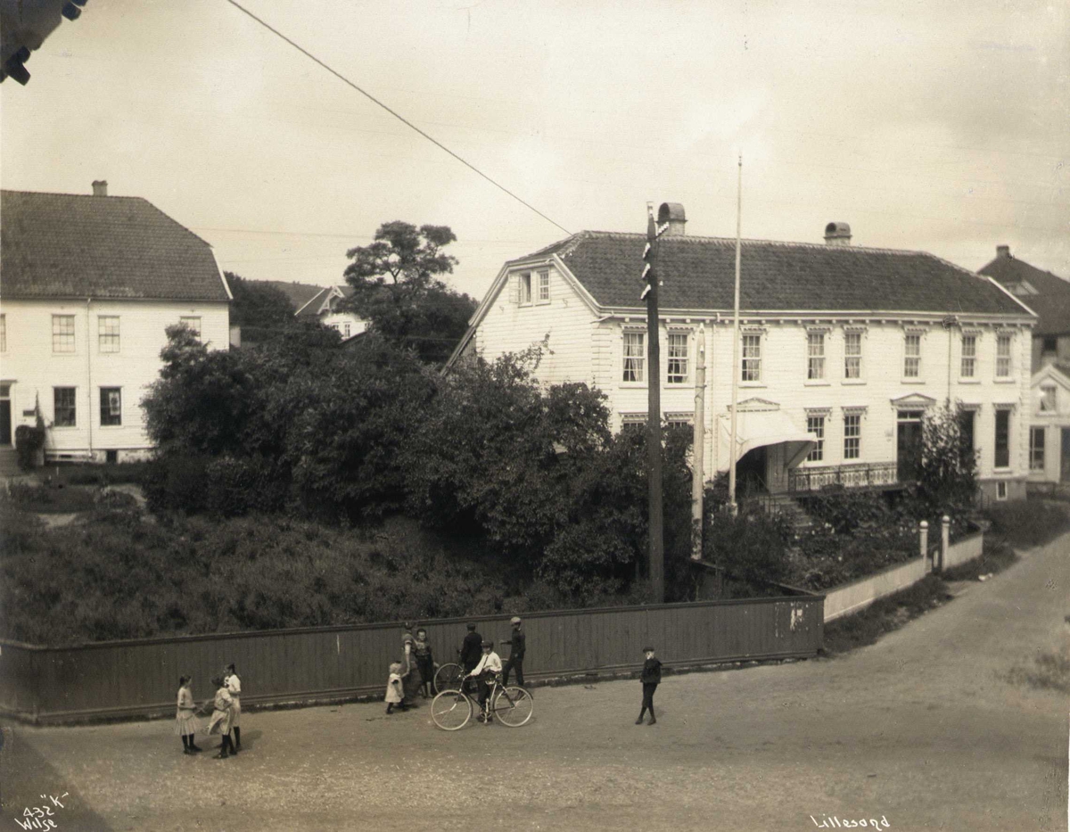 Gateparti med trebygninger, Lillesand, Aust-Agder. Barn leker i forgrunnen. Huset til høyre er Carl Knudsen-gården, til venstre ligger Henschien-gården (senere rådhus).