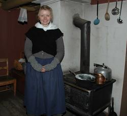 Kvinne i tidriktig kostyme foran vedkomfyren i et av husene 