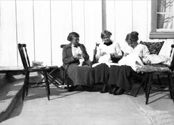 Tre kvinner sitter å driver med håndarbeid på en veranda. Ro