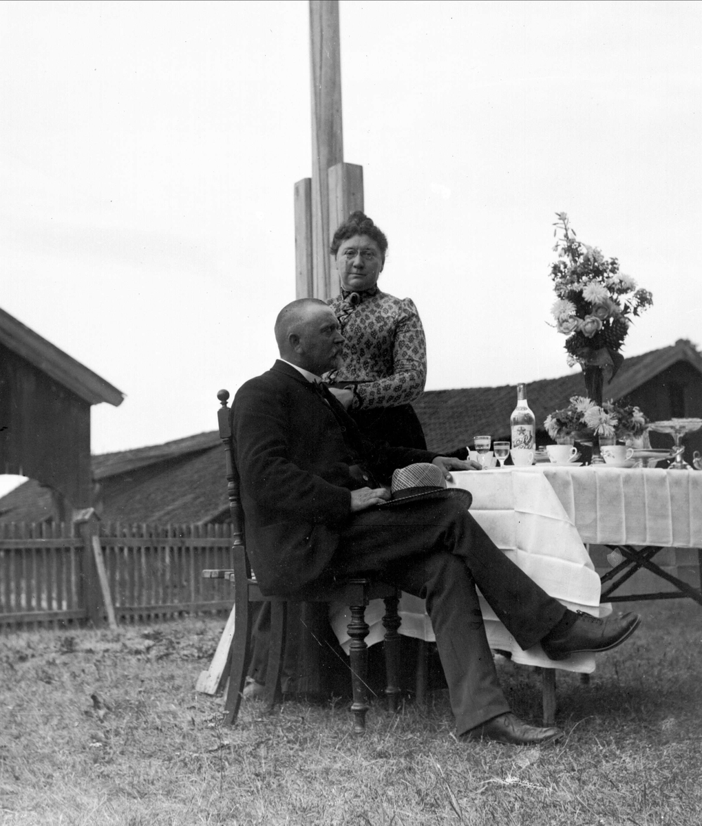Gruppeportrett, mann og kvinne ved bord i hagen. Vinhandler Larsen m/frue, fra Bygdøy.