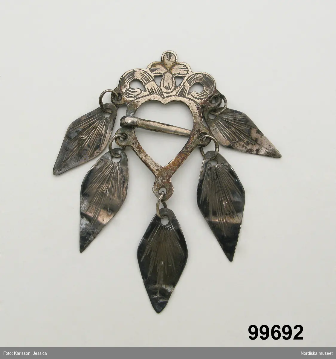 Hjärtformad sölja av silver med ornamenterat krön och 5 lövformade hängen, nål i mitten. Stämplar saknas.

Berit Eldvik maj 2006