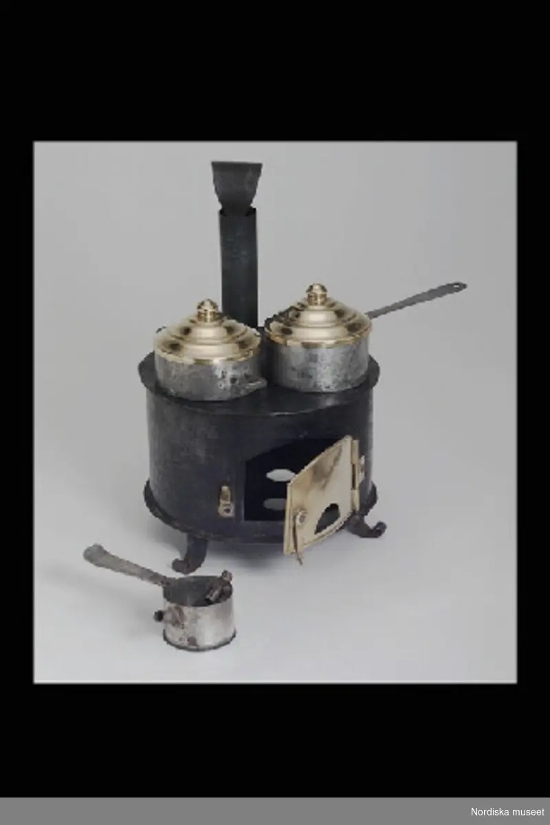 Inventering Sesam 1996-1999:
Diam  12,5   H med skorstensrör  19 (cm)
Dockskåpsspis, järnspis, med kokkärl (a-c), spis av svartlackerad bleckplåt, rund modell för två kokkärl på tre svängda fötter, öppningsbar eldstadslucka av mässing, löstagbart skorstensrör och brännare med handtag (a+), kastrull (b+) samt gryta med fläns (c+), båda av bleckplåt med mässingslock.
Tillhör dockskåp från omkr 1840, inv nr 145.505, inrett 1872 av leksakshandlare W A Bandel, Västerlånggatan, Stockholm, för dottern Thyra, vilken vid överlämnandet till museet själv gjort uppställningen av föremålen i dockskåpet. 
Anna Womack dec 1997