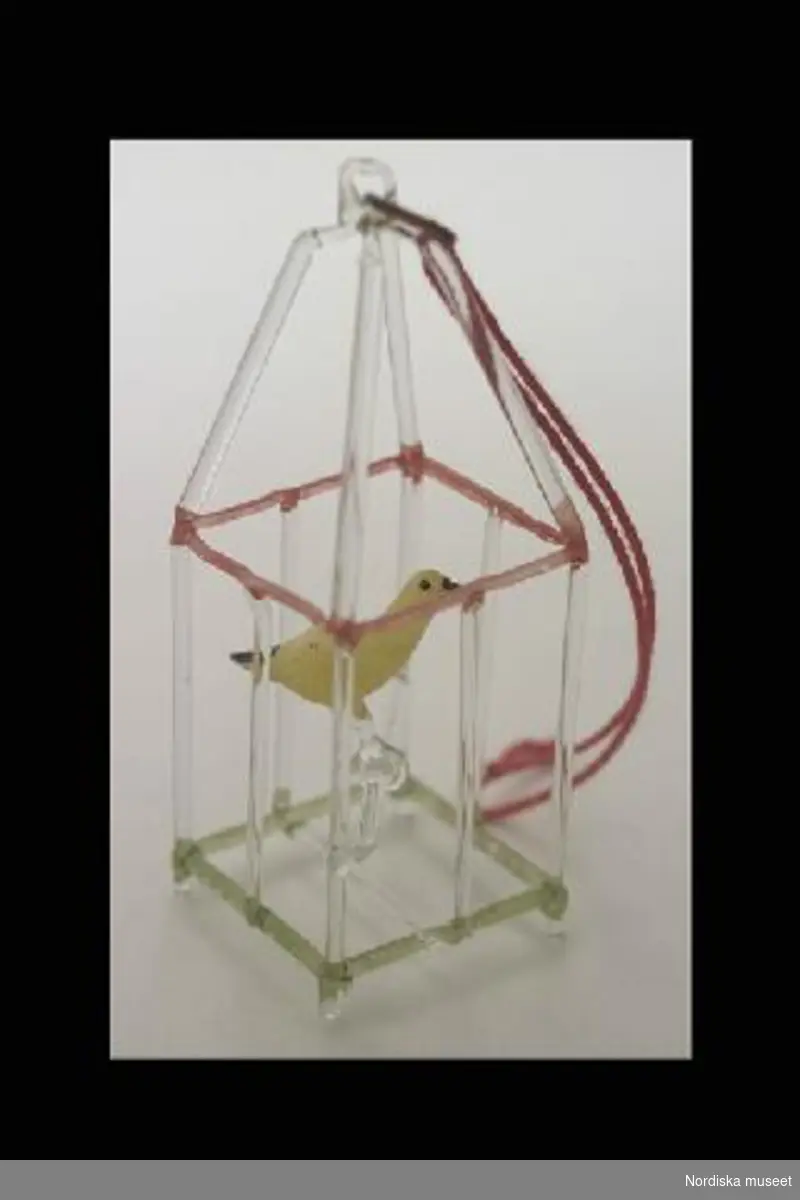 Inventering Sesam 1996-1999:
H 7,2  L 2,5   B 2,5 (cm) (bur)
a) Fågelbur med fågel, av glas, buren delvis färgad röd och grön,  med gul fågel på glaspinne. Glasögla med rött snöre.
b) Fågel av glas ( L 1,9 cm), vit kropp, gröna vingar och stjärt, i ryggen krok av gulmetall.
Brukad av Catarina (född 1919) och Anne-Sophie (född 1921) von Heidenstam, döttrar till givaren.
Tillhör dockskåp 220.405 med inv nr 220.406-220.760
Charlotta Dobson Hoffman okt 1997