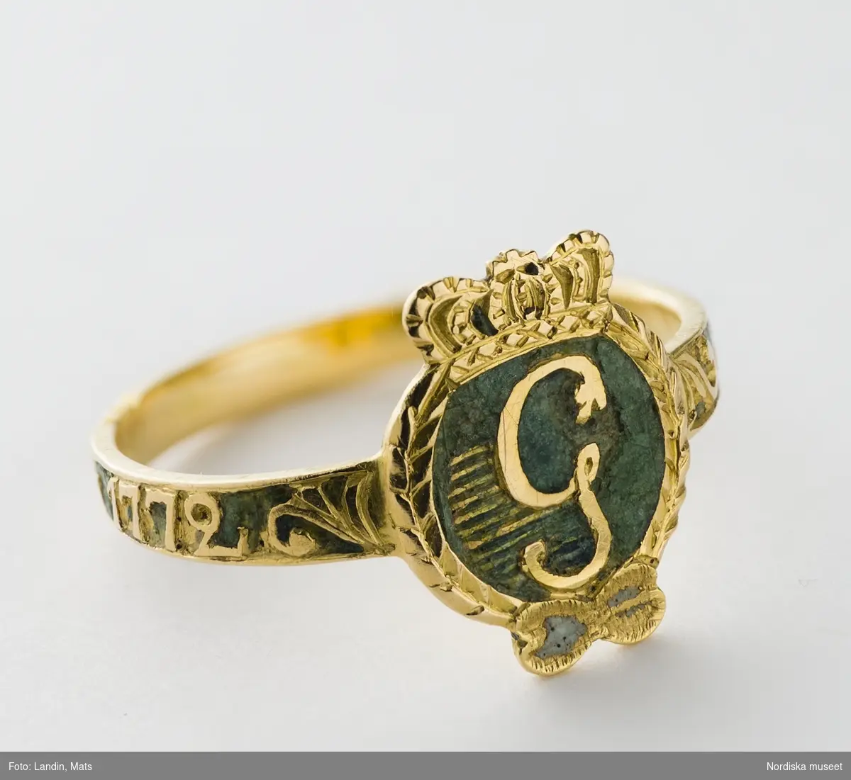 Ring av rött guld,  med Gustav III:s monogram i guld mot grön emaljbotten, s.k. revolutionsring. Runt skenan graverad inskription "den 19 aug 1772". 
/Ingrid Roos 2006-09-26