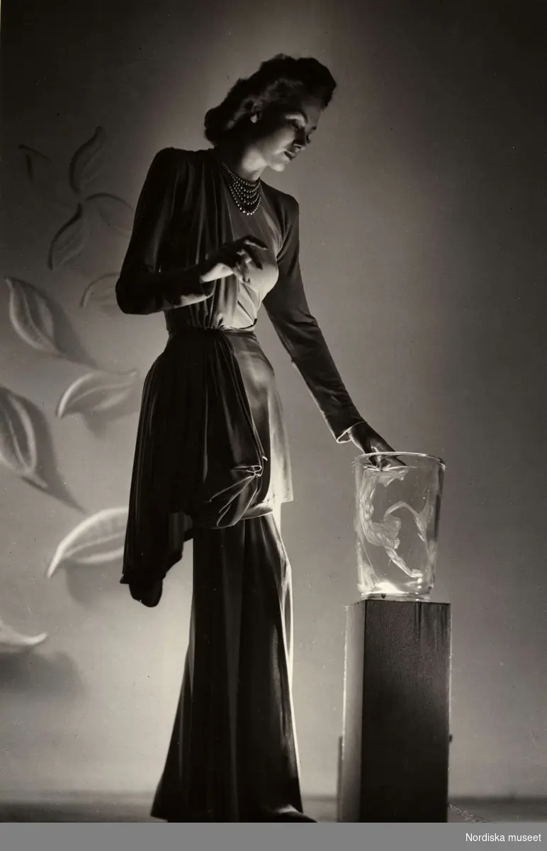 Franska avdelningen på Nordiska Kompaniet 1944. Kvinnlig modell poserar i dramatisk ljussättning iförd draperad aftonklänning. På ett podium intill henne en glasvas och på väggen bakom ett arrangemang med blad. 