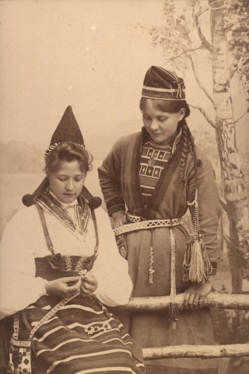 Ateljéporträtt. Två kvinnor i folkdräkt. Kvinnan till vänster bär en dräkt från Rättvik, Dalarna och kvinnan till höger bär en samisk dräkt.
