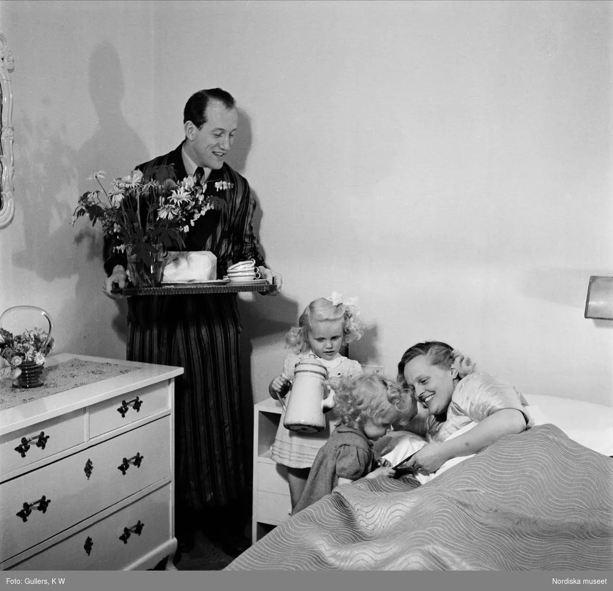 Mors dag. Två flickor och en man med kaffebricka uppvaktar mor vid sängen.