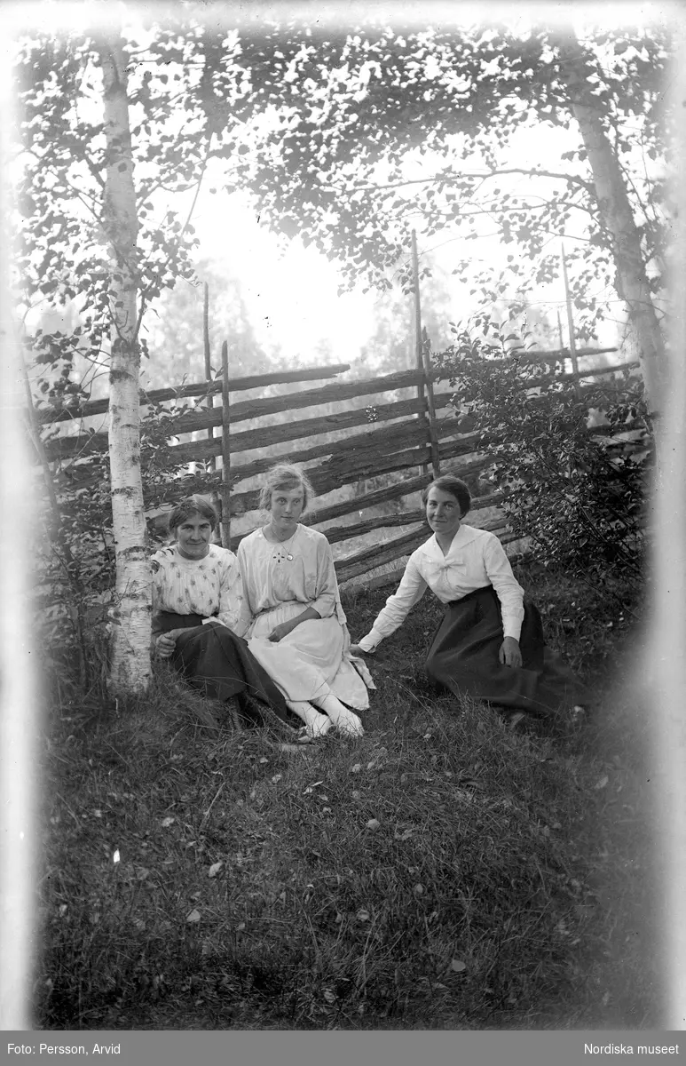Tre kvinnor sitter i gräset vid ett staket. Sörsjön, Transtrand sn, Dalarna.