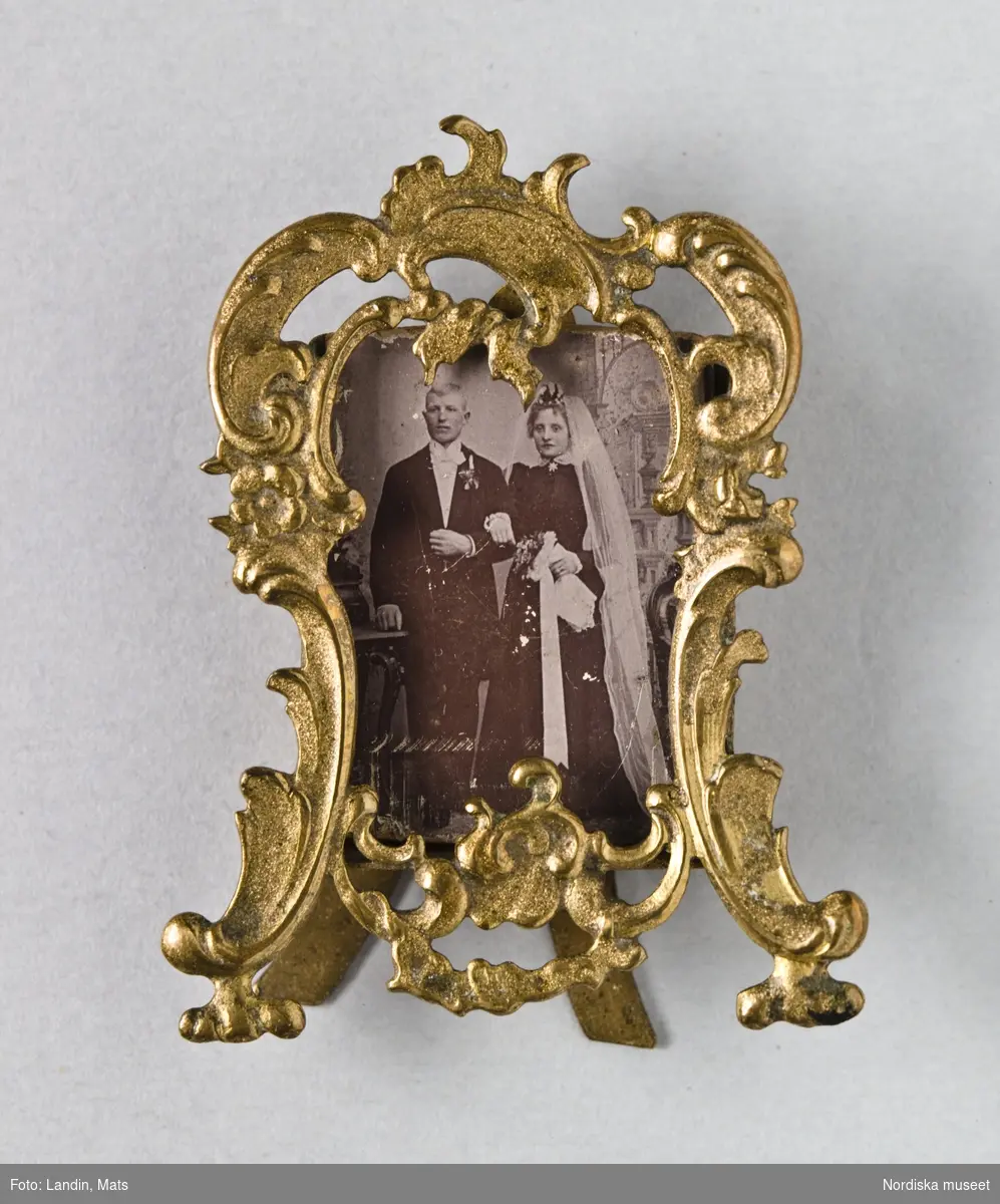 Bröllopsfoto med utsirad, genombruten ram i gulmetall. Föreställer Hilma och Edvin Jonsson, gifta 1895. Hon i svart brudklänning med vit slöja
Nordiska museet inv.nr 285228A