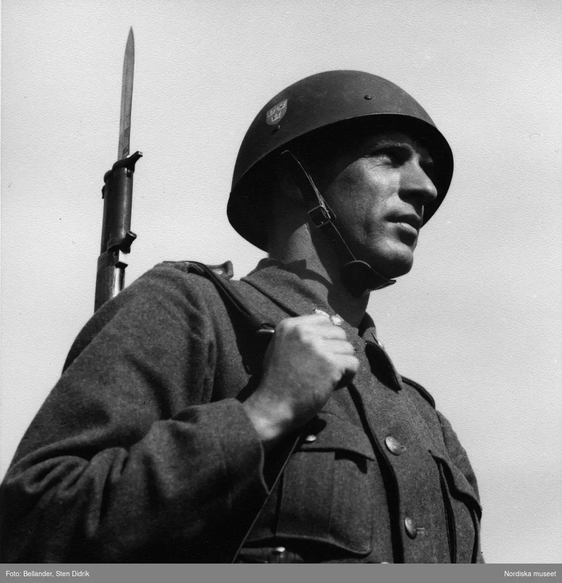 Svensk soldat i uniform och hjälm. Han bär sitt gevär med bajonett över axeln.