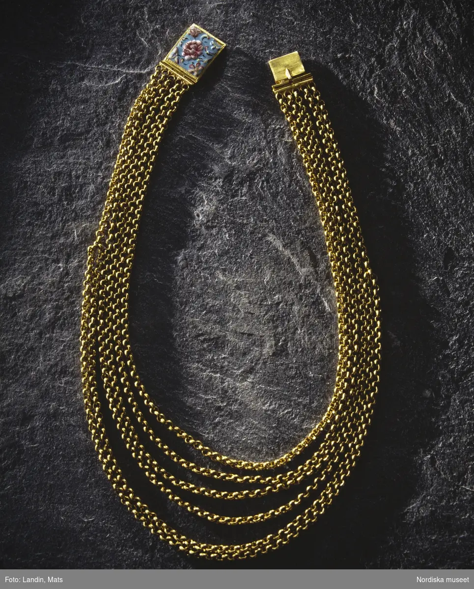 Halskedja av guld med lås i emalj. Låsets baksida försett med Kung Karl XI:s namnschiffer. Föremål ur Nordiska museets samlingar invnr:
219188.
