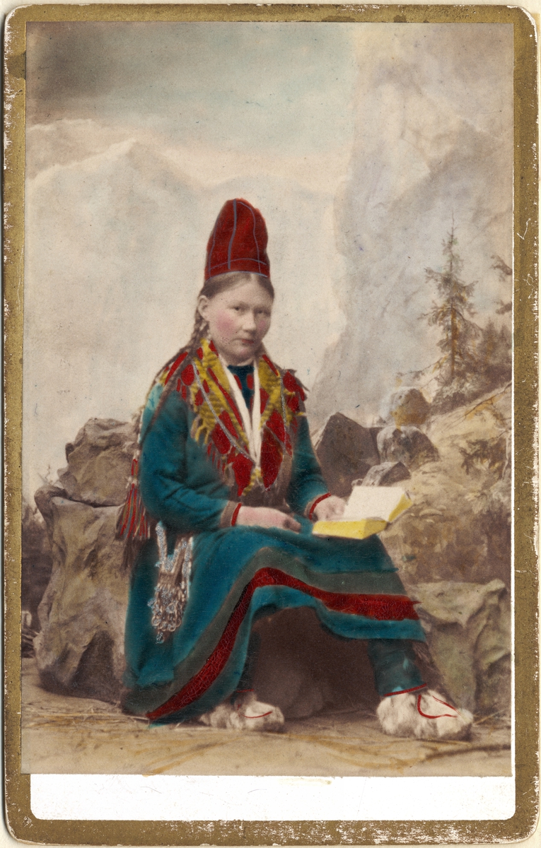 Ateljébild. Porträtt av kvinna i samisk dräkt med uppslagen bok. Handkolorerat foto.