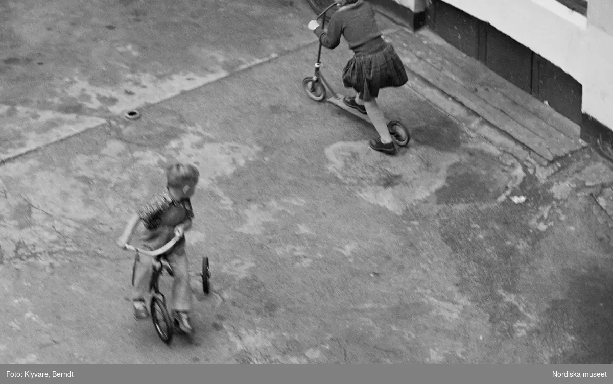 Barn leker på gård. En pojke på en trehjuling och en flicka på en sparkcykel.