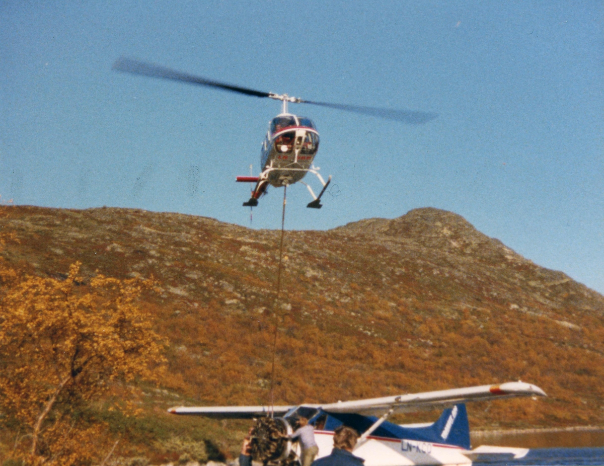 Ett helikopter i lufta over et fly på bakken. Motoren til flyet hanger under helikopteret