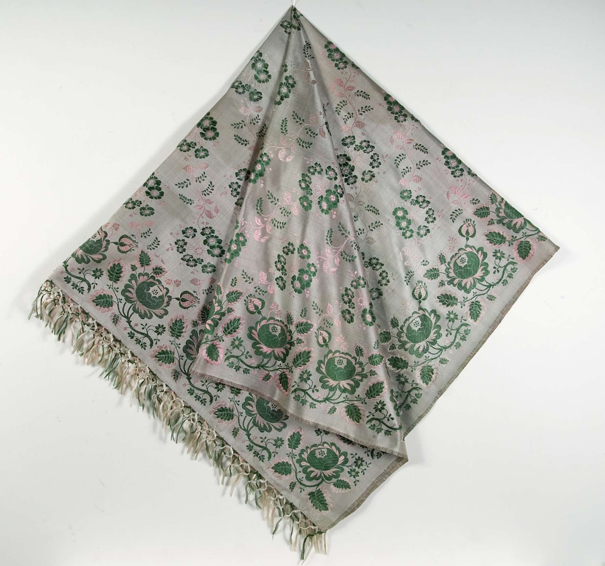 Kläde av ljusgrått siden med invävt blommönster i rosa, grönt. Iknuten frans på varpsidorna. Stockholms hallstämpel 1842.