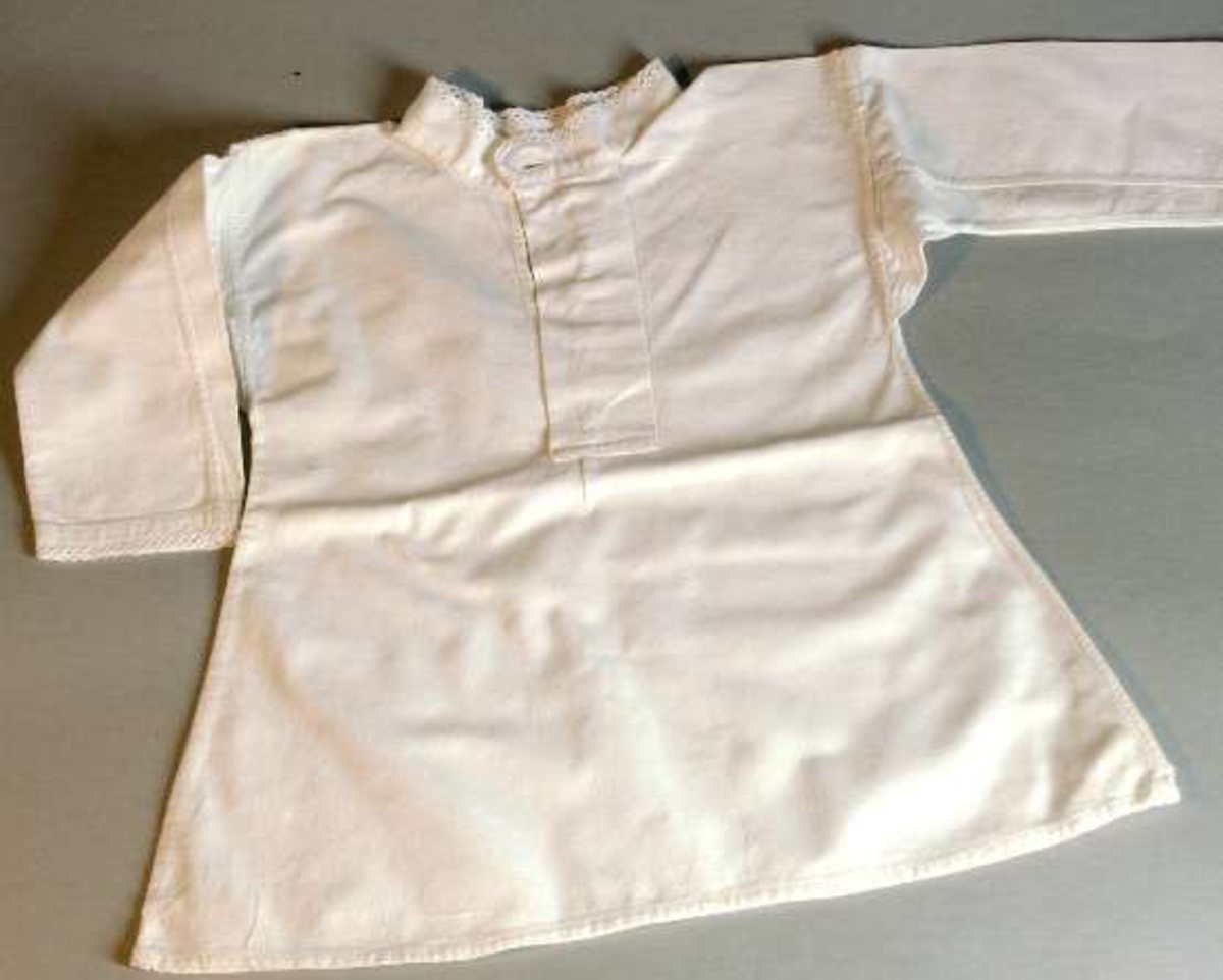 Skjorta av vit bomullslärft. Rund hals och ärmar genombrutet band av bomull. Knäpps fram med en knapp i ståndkragen. Fällsömmar. Maskinsydd.
