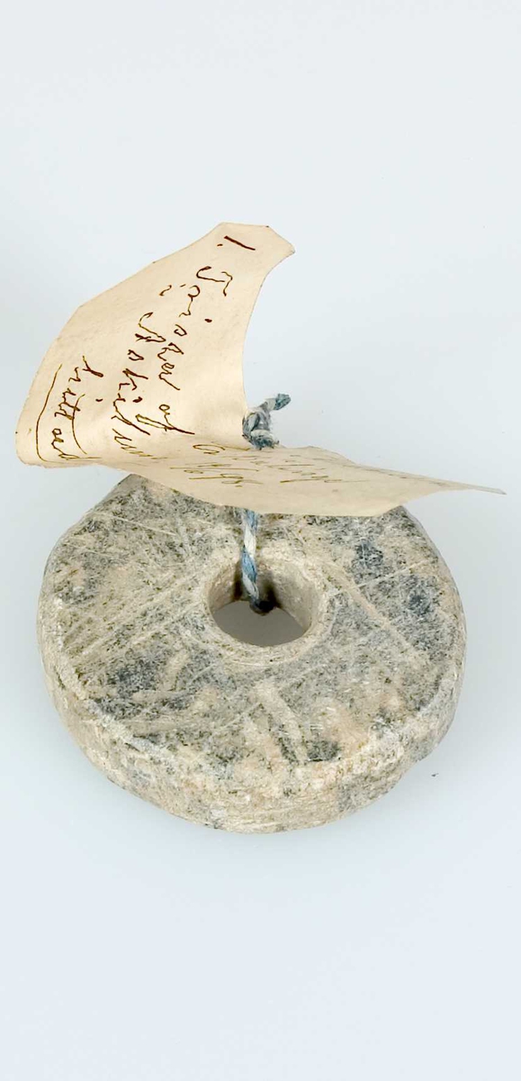Sländtrissa av sten. Etikett av papper fäst i trissan, handskriven text: 1. Trissa af Enkan Andersson i Äskilunda i Bälinge. Hittad där14/6 98.