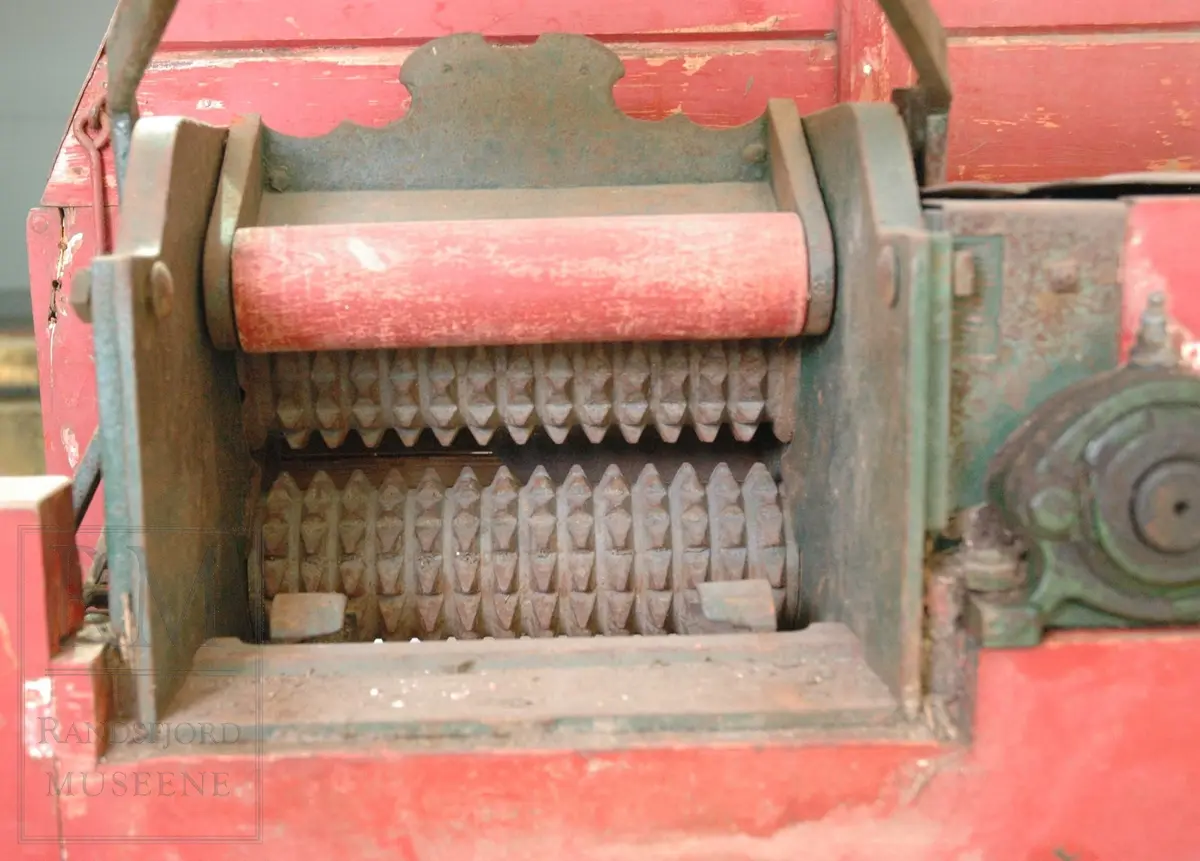 Rødmalt hakkelsmaskin i tre med tannhjul og kniver i jern. Svinghjulet mangler. 