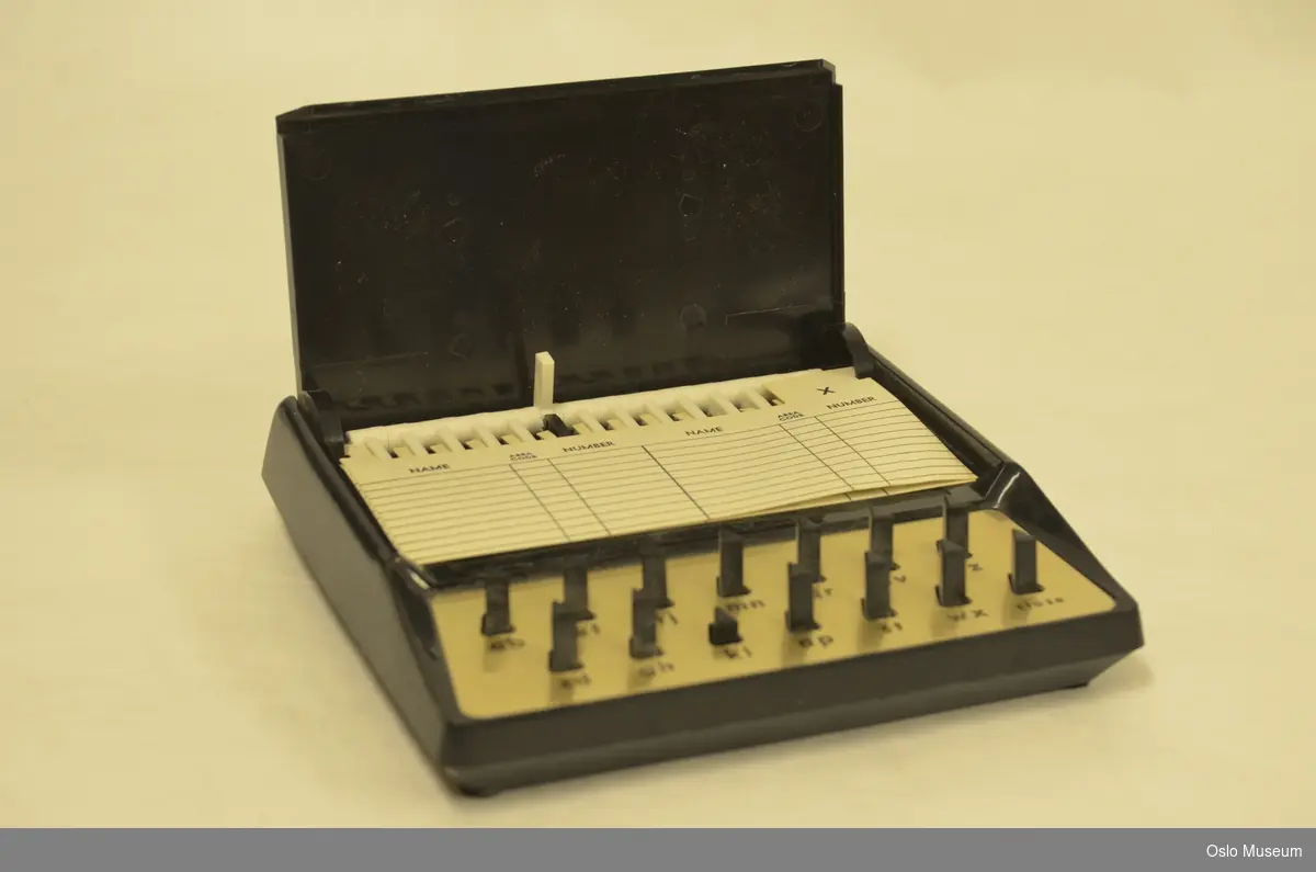 Telefonregister i sort og brun plast, metall og papir med bokstavtastatur. Lokket spretter opp ved å trykke valgt bokstav.