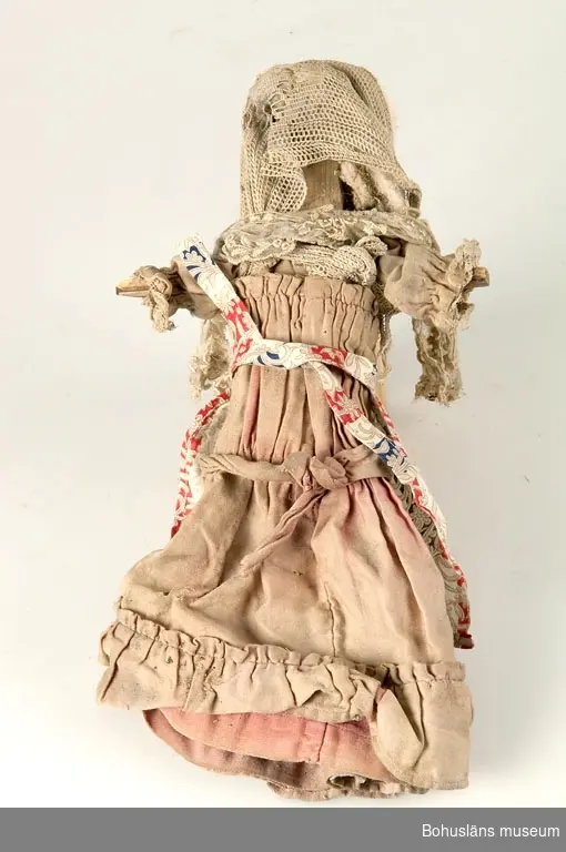 Hemmagjord trädocka från tidigt 1900-tal. Dockkroppen är grovt utskuren ur ett stycke furu och har ansikte med antydan till mun, näsa och ögon, påspikade armar som står rakt ut utan händer och raka ben utan fötter. Hår i form av bomull. Dockan är klädd i  en mycket blekt rosa klänning av sekelskiftessnitt med volang och garnering och med huvud- och axelschal av vit spets.
Klänningen verkar ha hört till en annan, finare dockkropp.

Enl. givaren kan dockan ha tillhört Ingeborg Hammar i Dragsmark,  f. 1916. Huset där dockan är funnen är byggt 1916, Klostergård 3, Dragsmark. 
Dockan är funnen i huset sommaren 2000 under en golvbräda på  vinden.