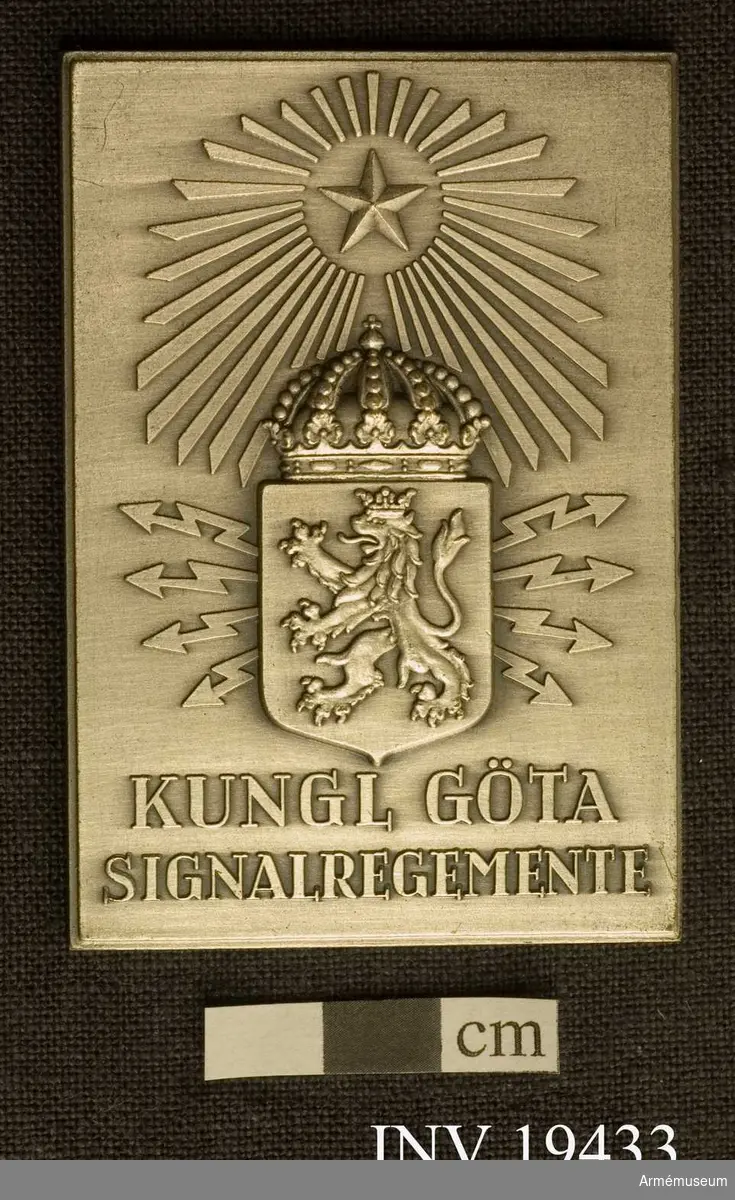 Grankvistemblemet. Plaketten är märkt på baksidan "Nr 3. H.Seitz 25/8 1962 Fästningsmuseum Karlsborg". På framsidan Kungliga Göta Signalregementes symbol: ett krönt lejon med blixtar och ovanför en femuddig stjärna med strålar.