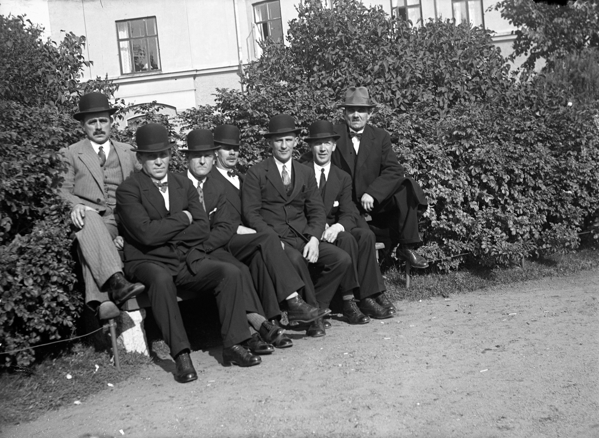 Sju män, alla klädda i kostym och hatt, sitter på en bänk framför en plantering vid ett bostadshus.